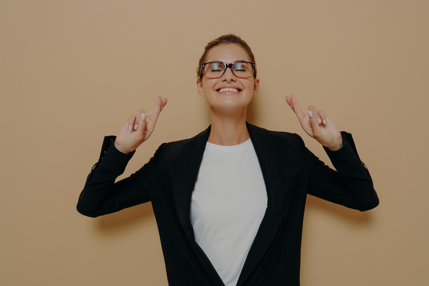 jonge hoopvolle vrouw die een bril draagt in een zwarte blazer over een witte t-shirt die haar vingers kruist met hoop foto