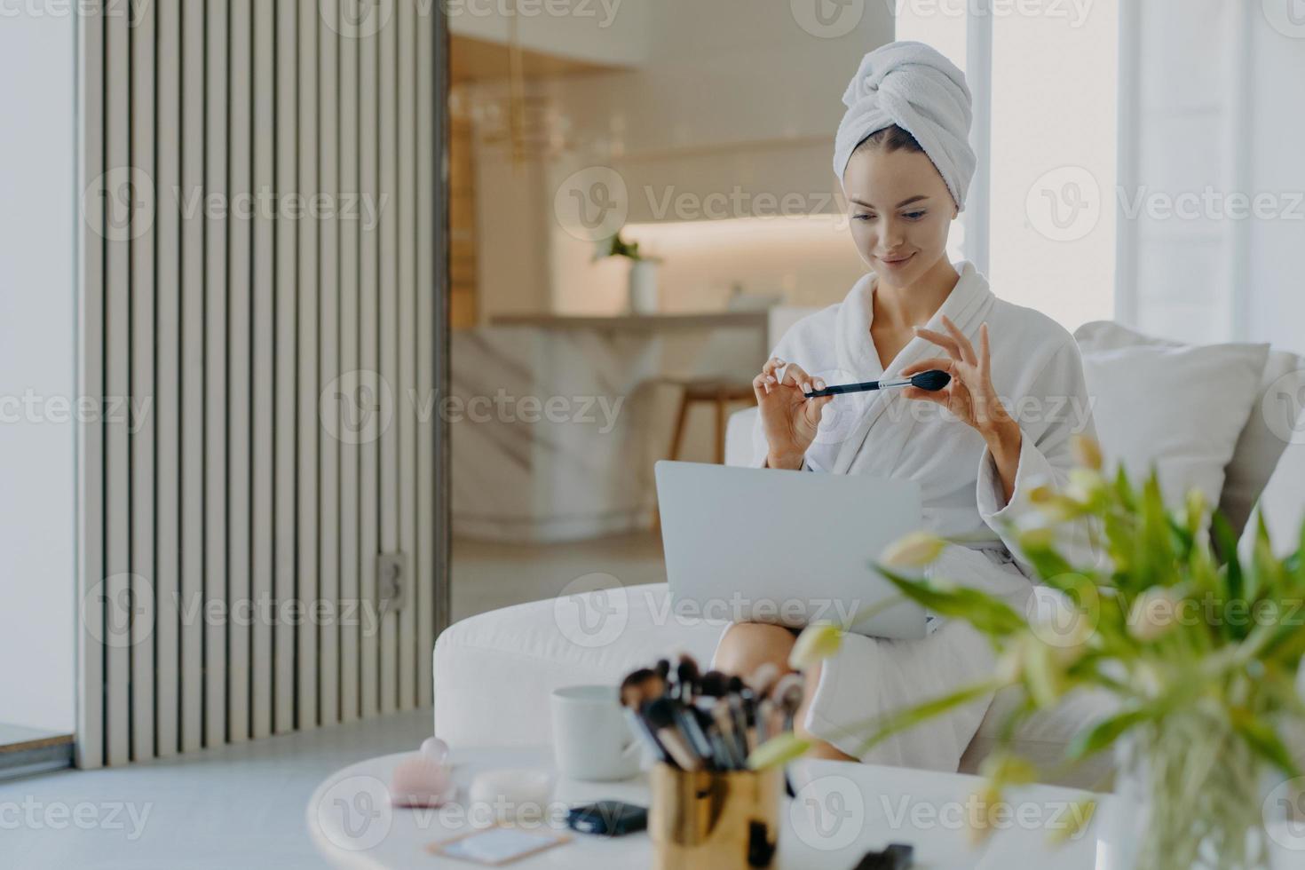 mooie jonge blanke vrouw draagt badjas en handdoek op hoofd houdt cosmetische borstel voor laptop computer geeft tutorial les hoe make-up te doen zit op comfortabele bank in modern appartement foto