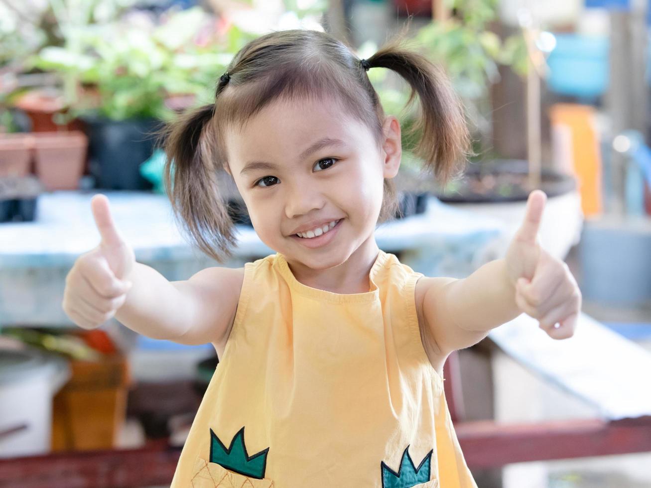 positief charmant 4 jaren oud schattig baby Aziatisch meisje, weinig kleuter kind met aanbiddelijk vlechten haar- glimlachen op zoek naar de camera tonen duimen omhoog gebaar Aan beide handen. foto