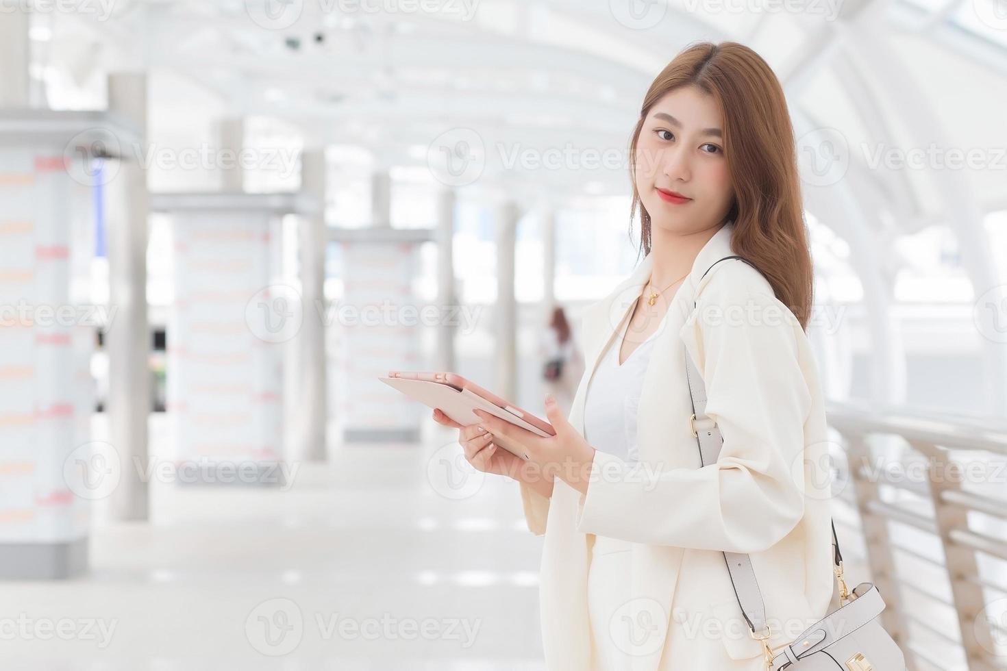 mooie jonge Aziatische professionele werkende zakenvrouw in wit pak is buiten om een klant te ontmoeten op kantoor haar hand tablet met zakelijke gebouwen en stad op de achtergrond. foto