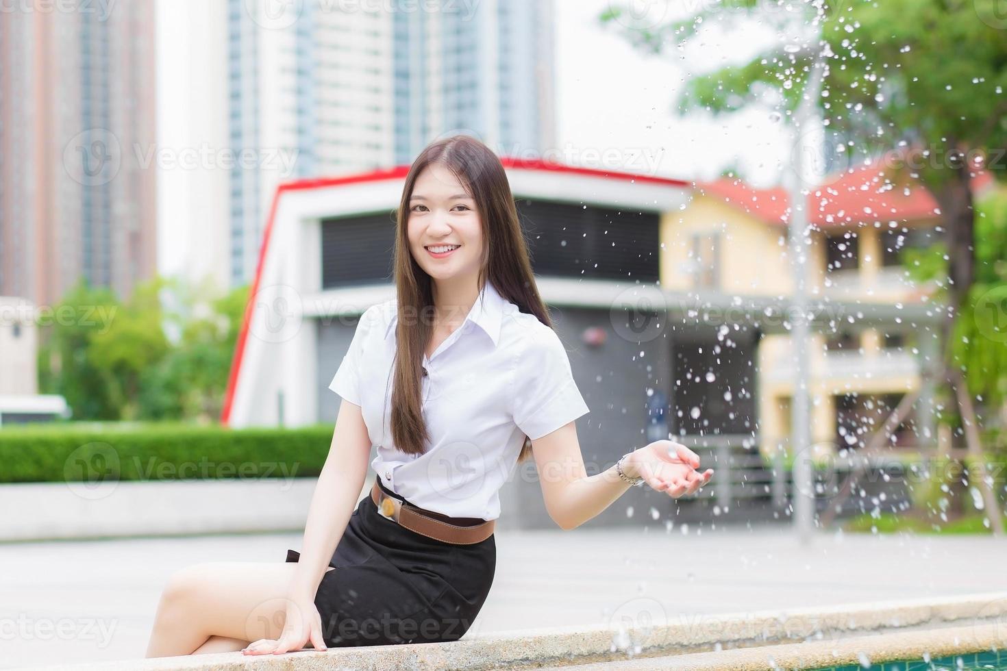 portret van volwassen Thais leerling in Universiteit leerling uniform. Aziatisch mooi meisje zittend schudden de water en glimlachen gelukkig Bij buitenshuis Universiteit. foto