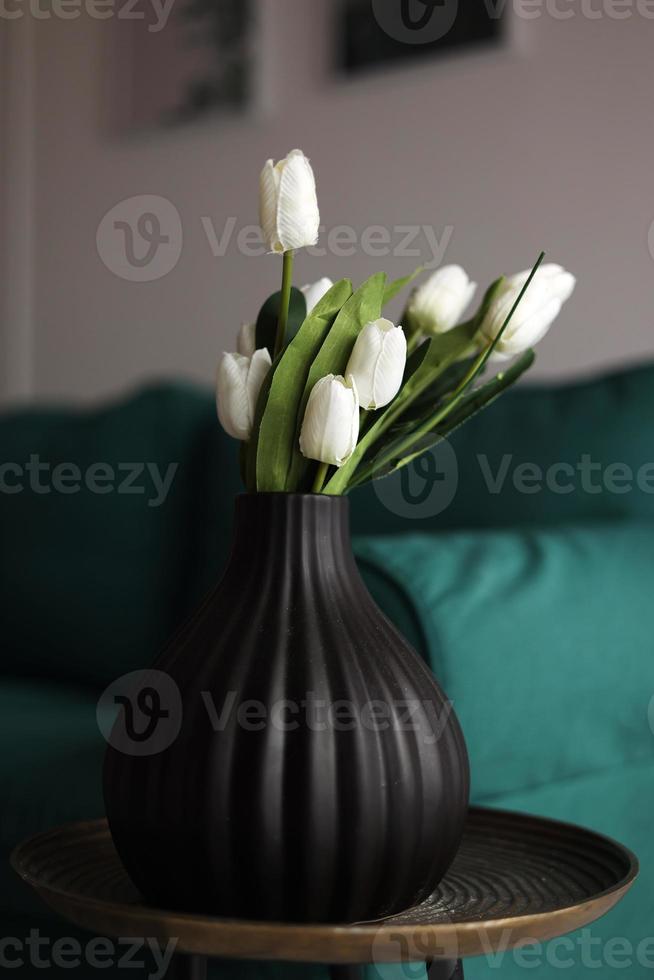 kunstmatig wit tulpen en groen bladeren in een zwart vaas Aan de tafel in huis interieur. foto