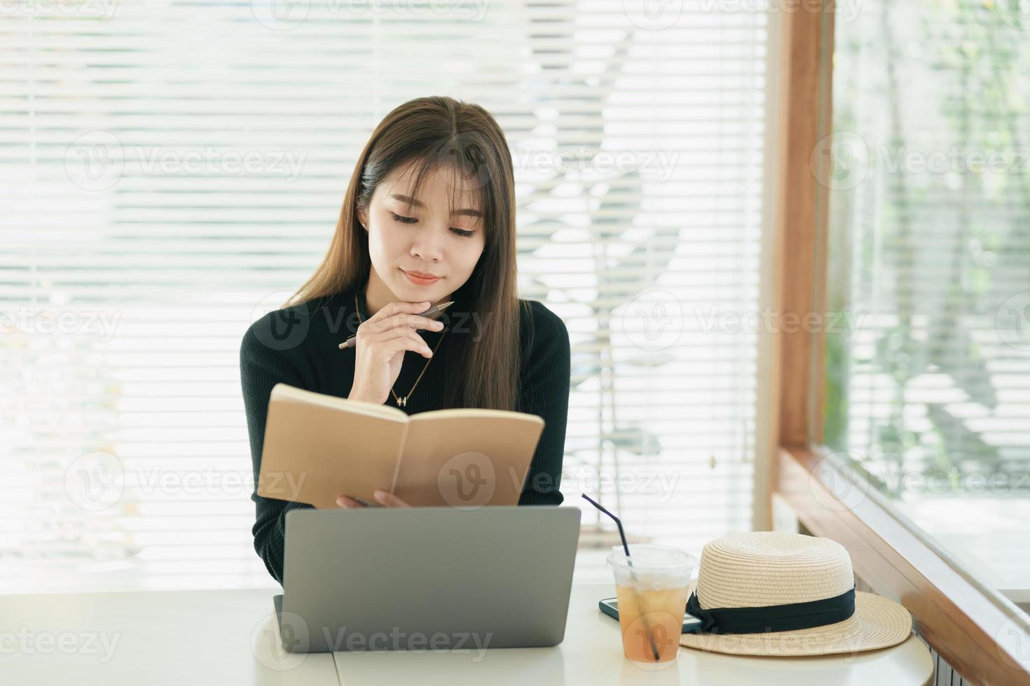 Aziatisch freelance vrouw glimlachen gebruik makend van pen maken mededelingen haar notitieboekje en werken Aan laptop Aan tafel Bij huis. ondernemer vrouw werken voor haar bedrijf Bij cafe. bedrijf werk Bij koffie winkel concept foto