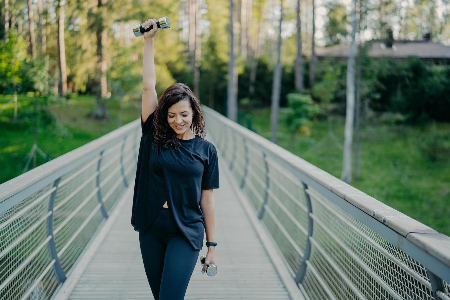 actieve jonge brunette sportvrouw oefent met halters, doet fitnessoefeningen buiten, gekleed in actieve kleding, ademt frisse lucht en werkt op spieren. vrouwen, kracht en bodybuilding concept foto