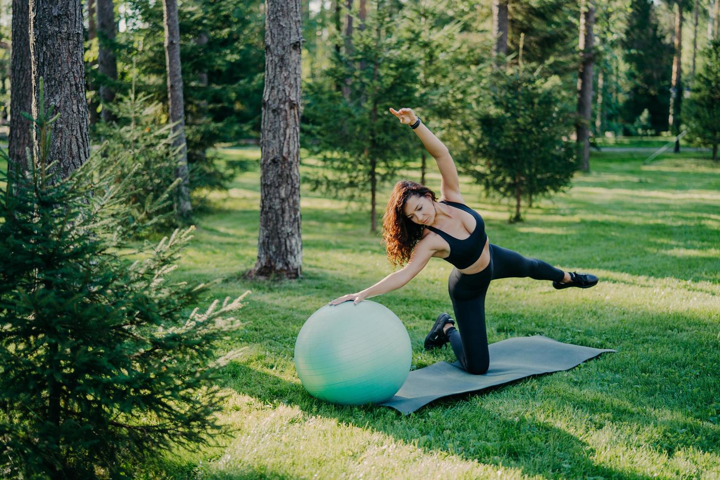 fitness, sport en gezonde levensstijl concept. buiten schot van slanke brunette Europese vrouw doet pilates en balans oefening met fit bal kantelt opzij poses in groen bos op gras bij karemat. foto