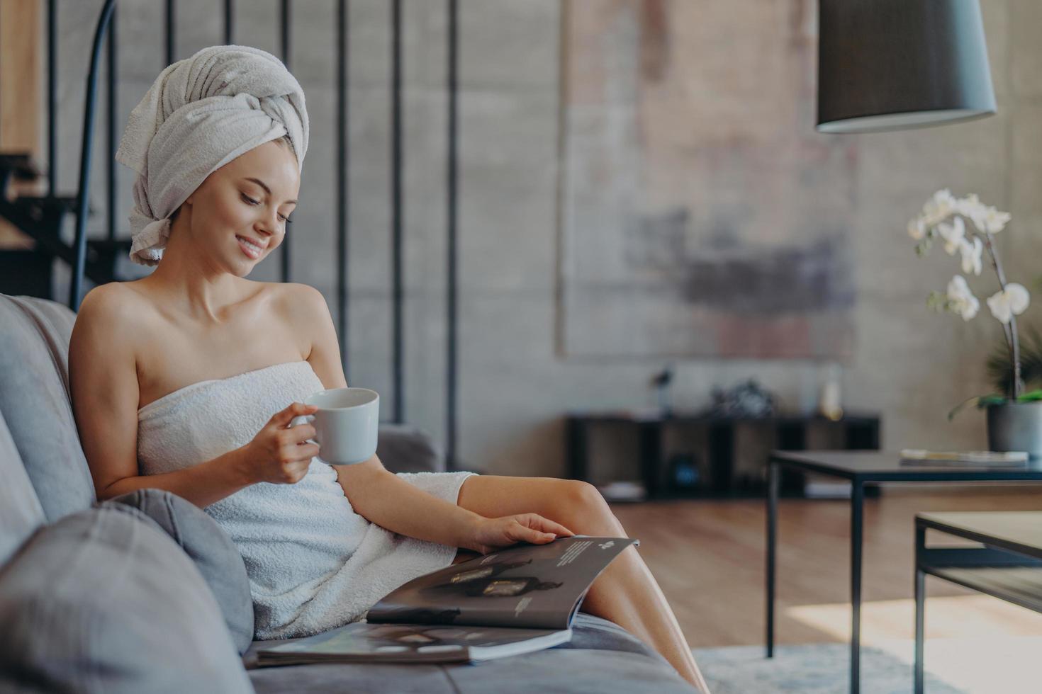 ontspannen lachende jonge vrouw gewikkeld in een handdoek na het douchen, drinkt koffie en leest beautymagazine, zit op een comfortabele bank tegen een gezellig interieur. wellness- en schoonheidsconcept foto