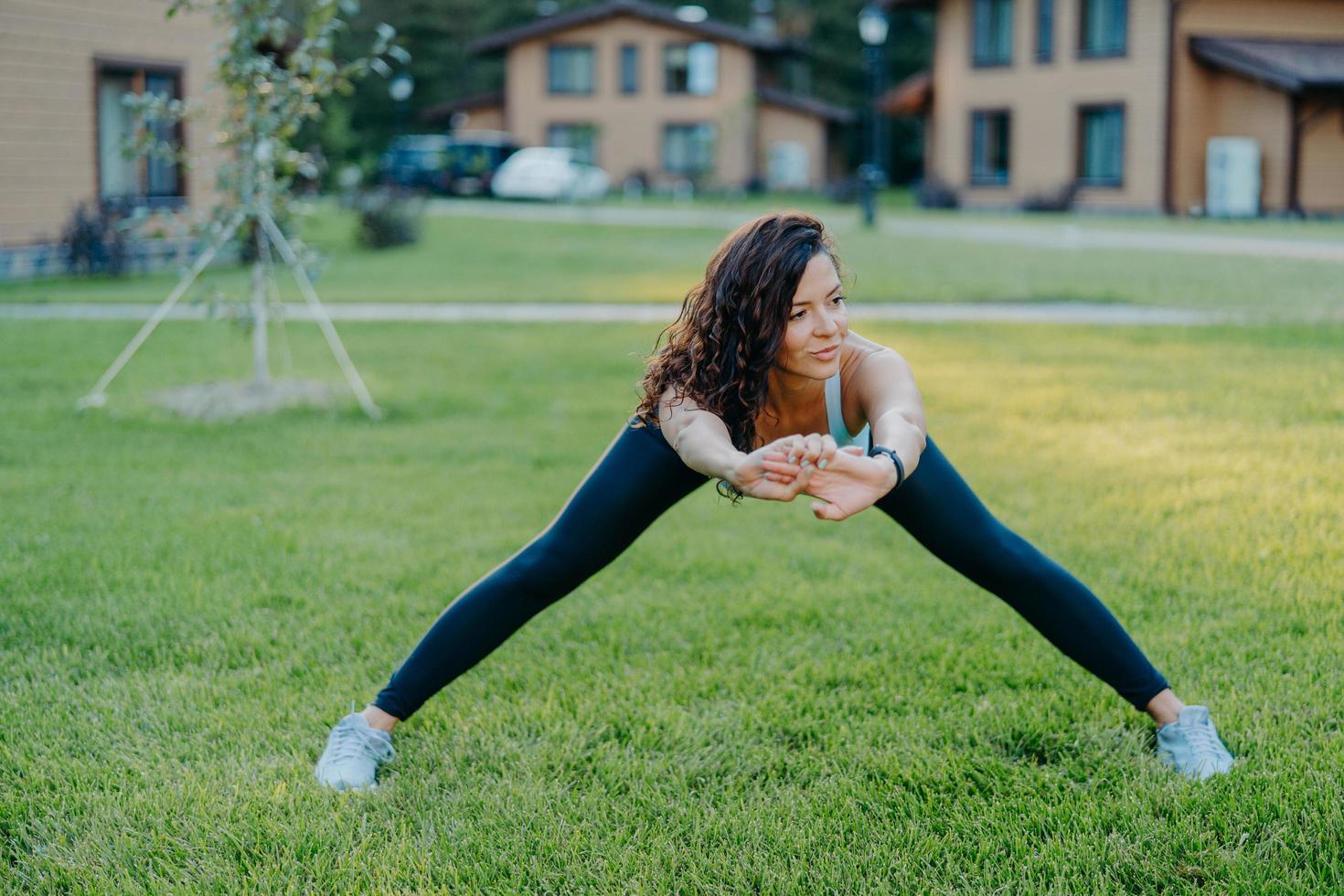 zelfverzekerde atletische vrouw demonstreert haar flexibiliteit, strekt zich buiten uit, doet oefeningen vroeg in de ochtend op groen gras in de buurt van huis leidt actieve levensstijl gekleed in actieve kleding blijft gezond foto