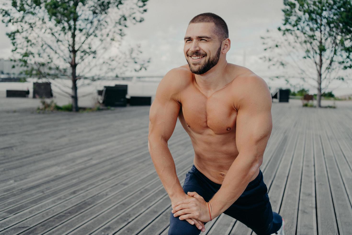 dagelijkse ochtendroutine en sportoefeningen concept. sportieve ongeschoren man bodybuilder met vrolijke uitdrukking doet rekoefeningen voor de benen, poseert met naakte torso, heeft training buiten. foto