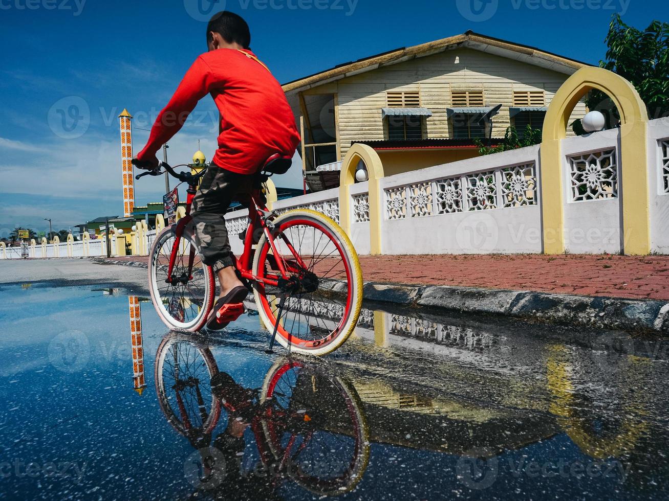 een jongen in rood rijden de fiets in de water plas Aan zijn manier naar de moskee foto