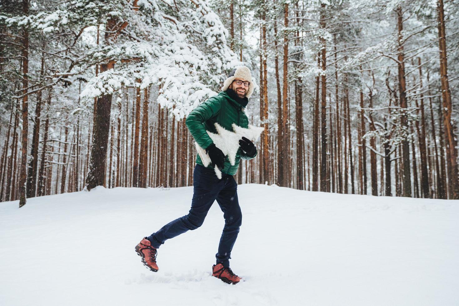 optimistische jonge man gekleed in warme winterkleren, heeft plezier buiten in winterbos, adem frisse lucht in, blij om veel sneeuw te zien, houdt atrficial dennenboom in handen. mensen, seizoen, weerconcept foto