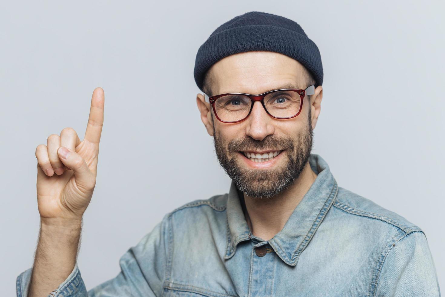 Positieve knappe bebaarde man steekt voorwaartse fimger op, kijkt met vrolijke uitdrukking direct in de camera, draagt modieuze hoed, shirt, steekt voorvinger op als hij zich iets herinnert, modellen binnen foto