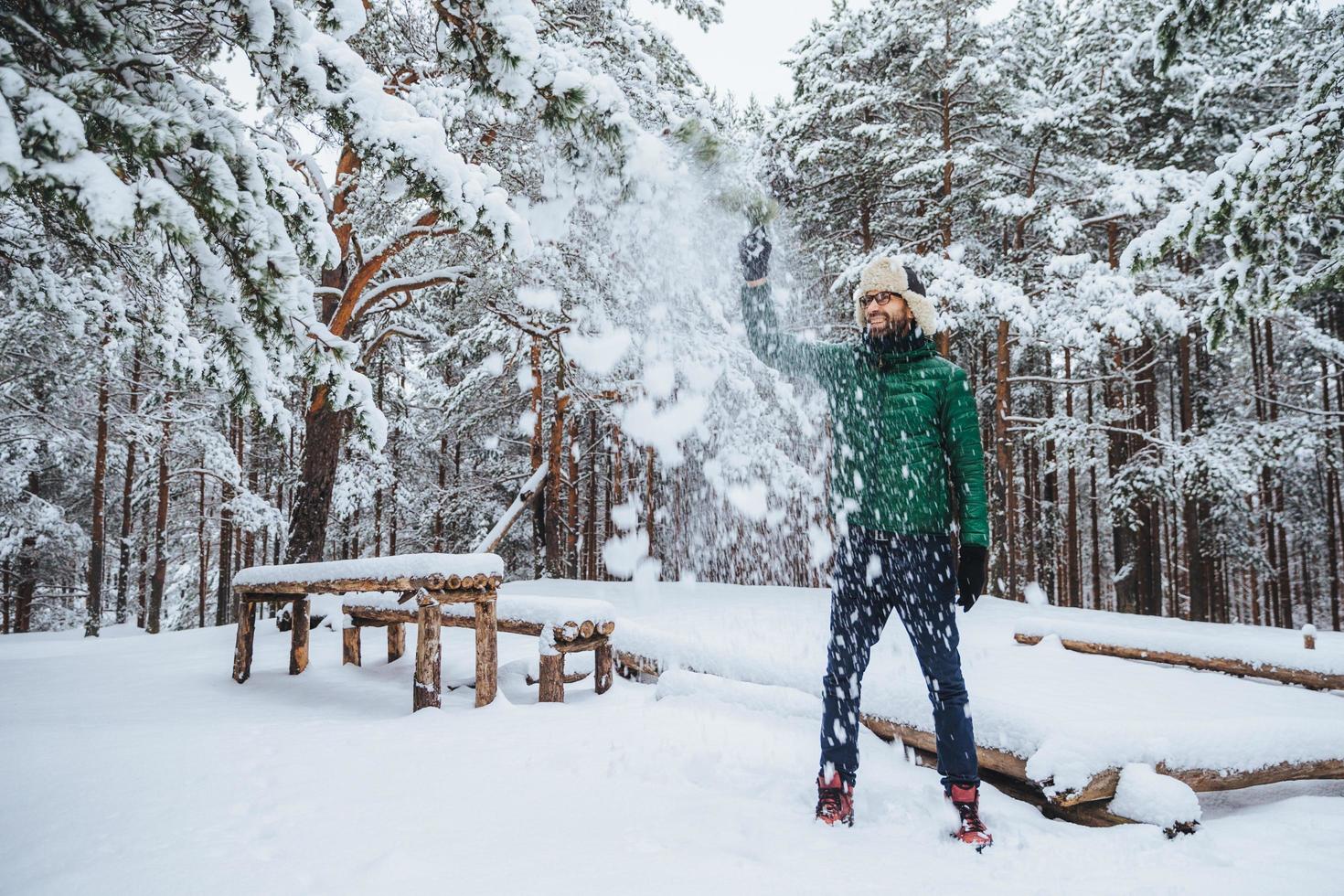 buitenopname van een knappe, bebaarde man gekleed in warme kleding, heeft plezier als sneeuw in de lucht wordt gegooid, brengt vakantie door in het winterbos, drukt positiviteit uit. recreatie en weer concept foto