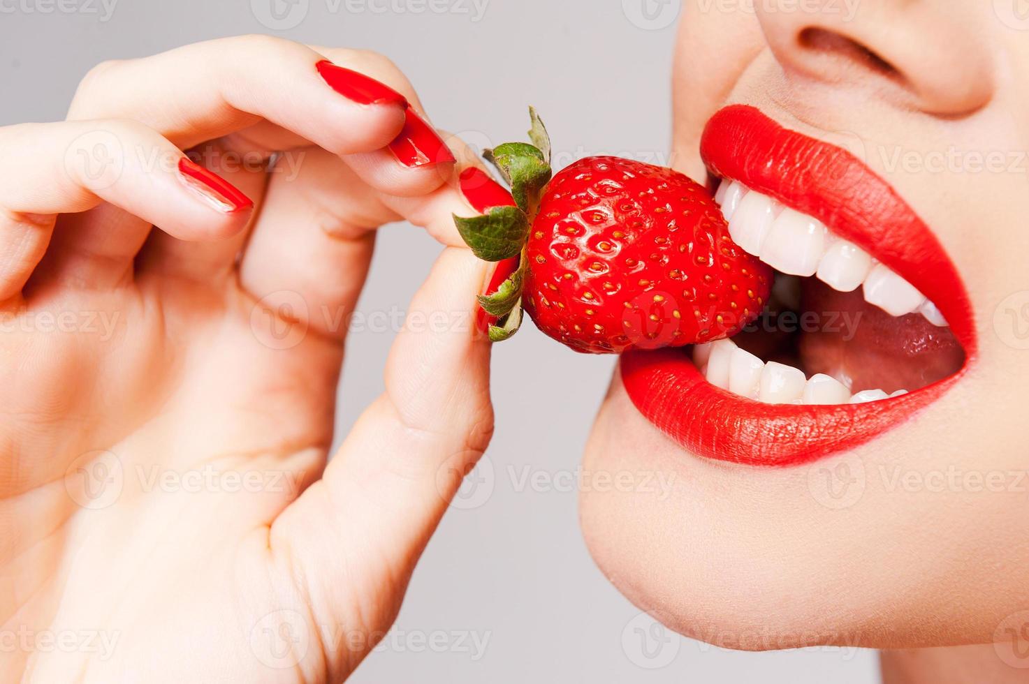 versheid komt in rood. detailopname van mooi vrouw Holding aardbei in haar hand- en proeverij het terwijl staand tegen grijs achtergrond foto