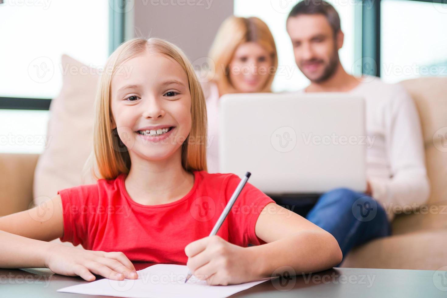 ik liefde mijn familie gelukkig weinig meisje tekening iets Aan papier en glimlachen terwijl haar ouders zittend in de achtergrond met laptop foto