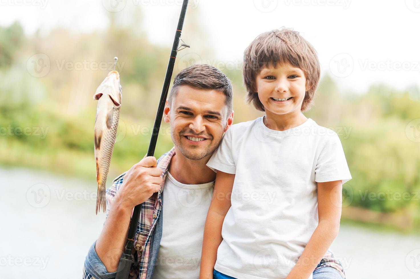 wij hebben gevangen het samen vader en zoon op zoek Bij camera en glimlachen terwijl Mens Holding visvangst hengel met groot vis Aan de haak foto