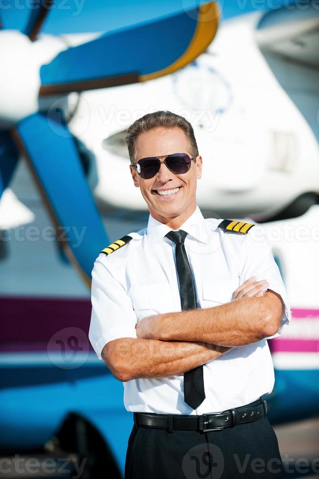 mijn baan is mijn passie. zelfverzekerd mannetje piloot in uniform houden armen gekruiste en glimlachen terwijl staand in voorkant van de vliegtuig foto