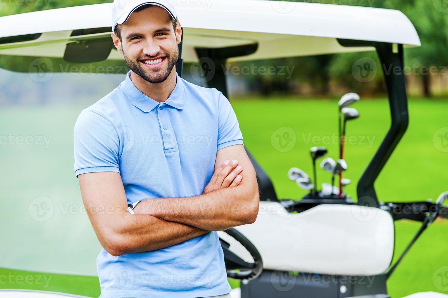 golf is mijn favoriete spel knap jong Mens houden armen gekruiste en glimlachen terwijl leunend Bij de golf kar terwijl staand Aan golf Cursus foto
