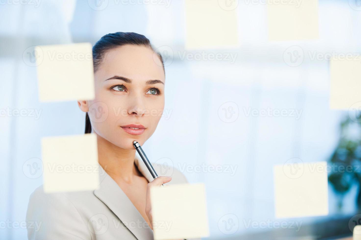 planning bedrijf strategie. attent jong zakenvrouw aanraken haar kin met pen en op zoek Bij de Zelfklevend aantekeningen gehecht naar de glas foto