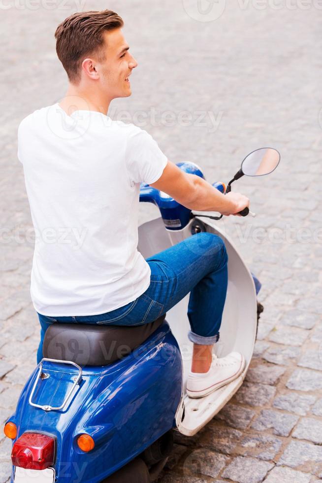 zorgeloos rijden. achterzijde visie van knap jong Mens in zonnebril rijden scooter langs de straat en glimlachen foto