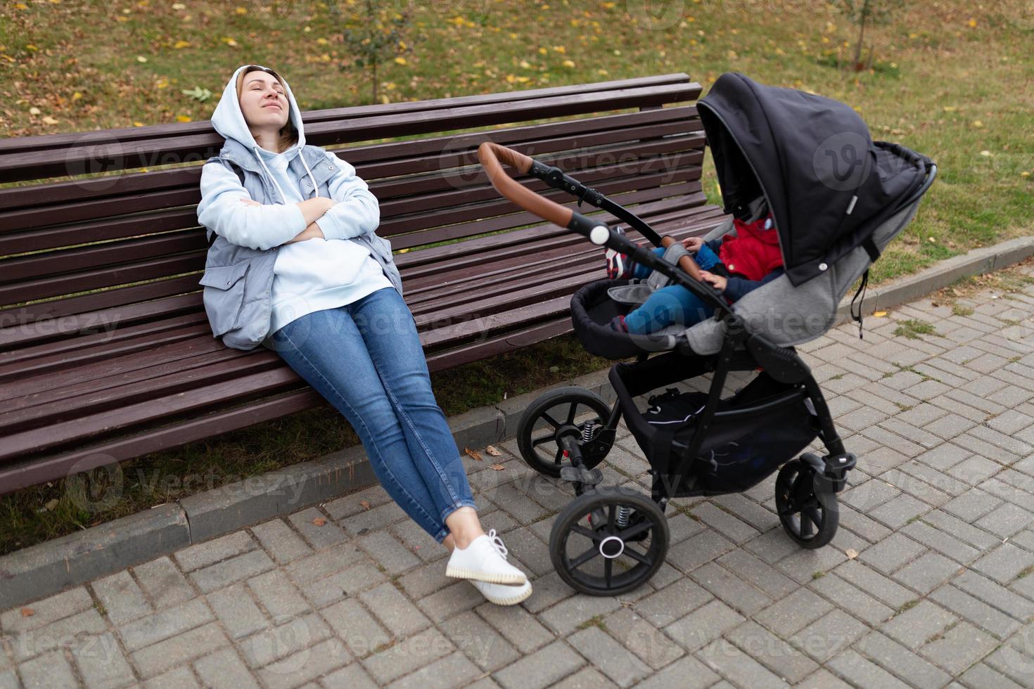 een jong vrouw slaapt Aan een bank De volgende naar de wandelwagen terwijl wandelen met haar klein kind foto