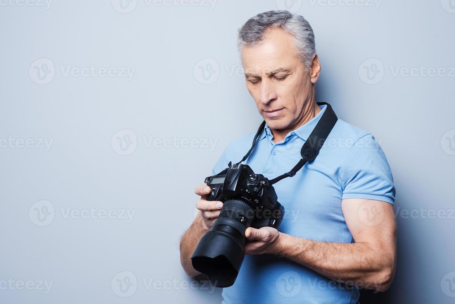 instelling de camera. portret van senior volwassen Mens in t-shirt Holding camera terwijl staand tegen grijs achtergrond foto