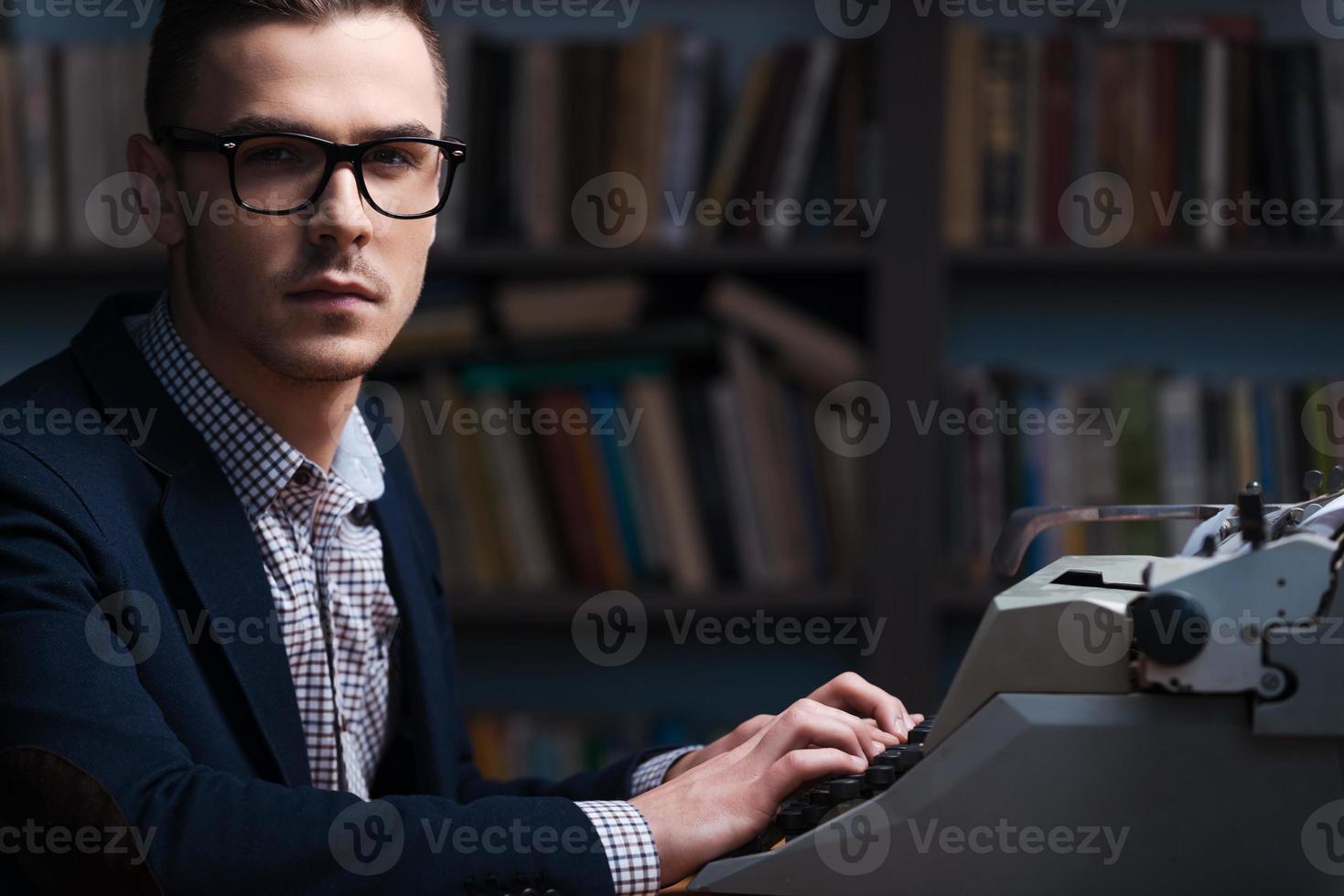 zelfverzekerd jong auteur. kant visie van zelfverzekerd jong auteur zittend Bij de schrijfmachine en op zoek Bij camera met boekenplank in de achtergrond foto