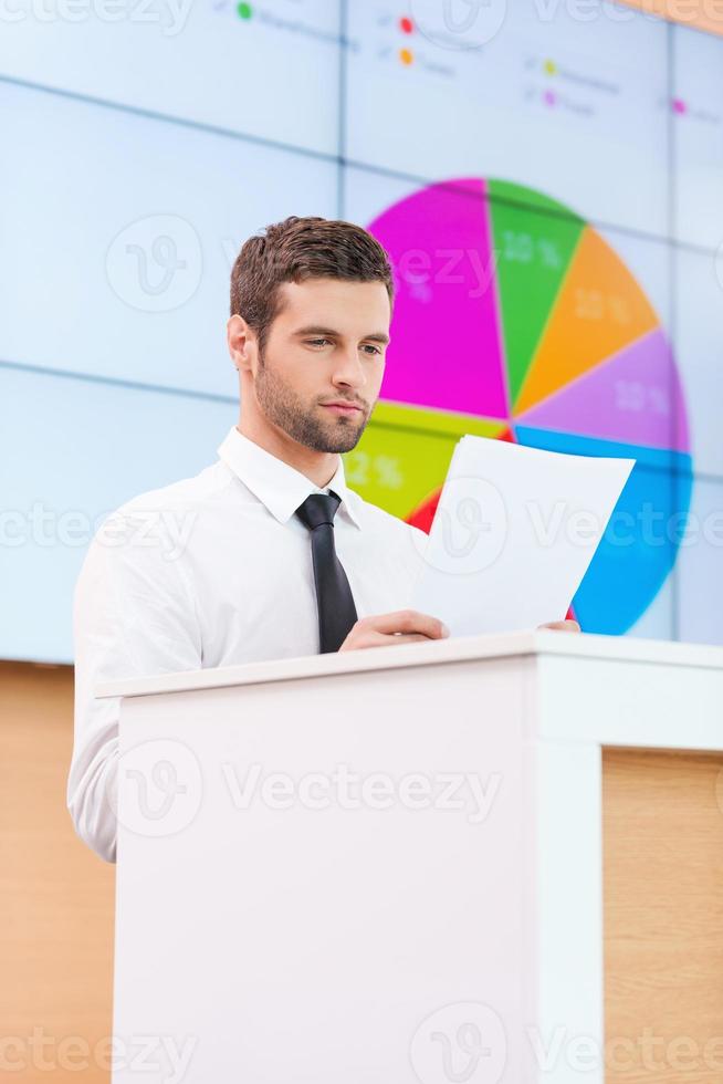 krijgen klaar naar presentatie. zelfverzekerd jong Mens in formele kleding staand Bij de tribune en op zoek Bij papier terwijl maken een presentatie met projectie scherm in de achtergrond foto