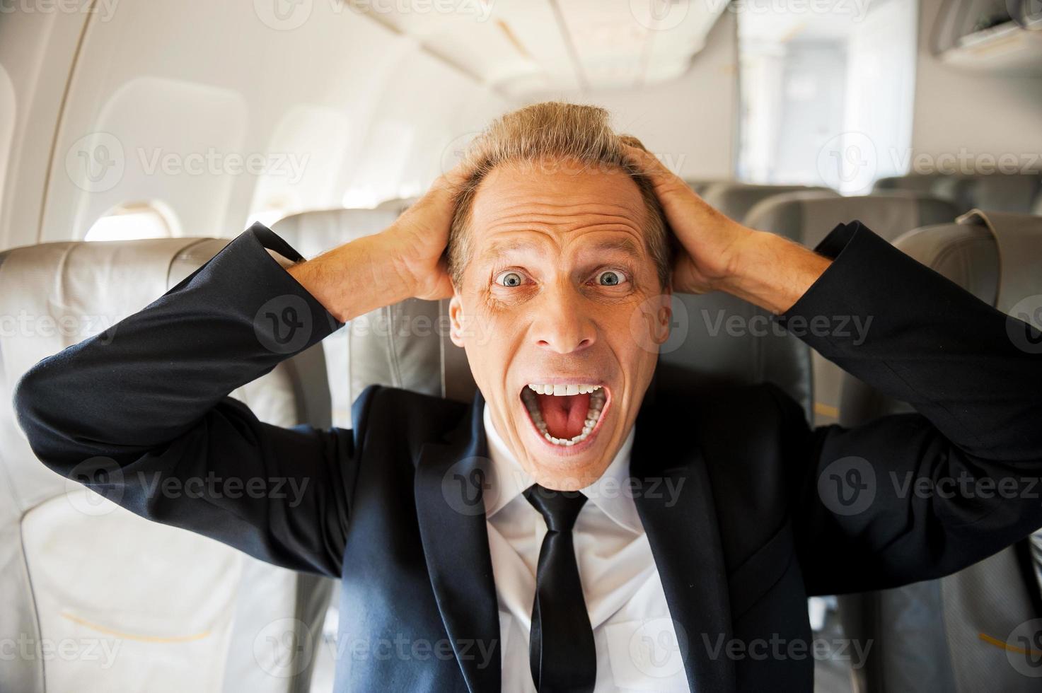 angst van vlucht. geschokt volwassen zakenman aanraken zijn hoofd met handen en op zoek Bij camera terwijl zittend Bij zijn stoel in vliegtuig foto
