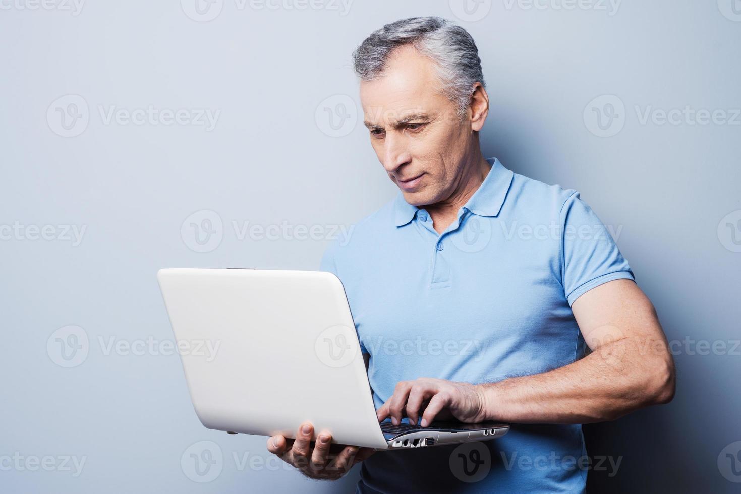 modern technologieën altijd Bij hand. vrolijk senior Mens in gewoontjes Holding laptop terwijl staand tegen grijs achtergrond foto