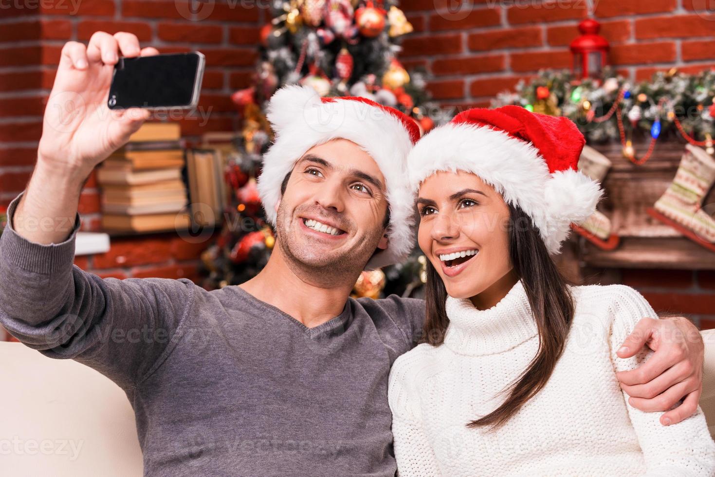 vastleggen gelukkig momenten. mooi jong liefhebbend paar bonding naar elk andere en glimlachen terwijl maken selfie met Kerstmis boom in de achtergrond foto