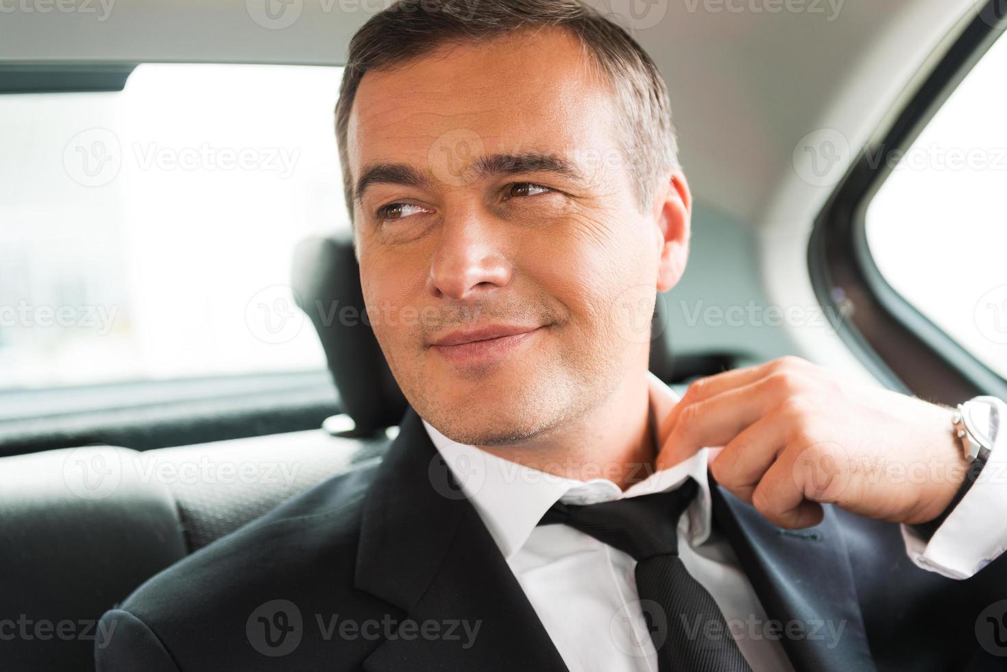 ontspannende na bedrijf reis. tevreden volwassen zakenman aanpassen zijn stropdas terwijl zittend Aan de terug stoel van een auto foto
