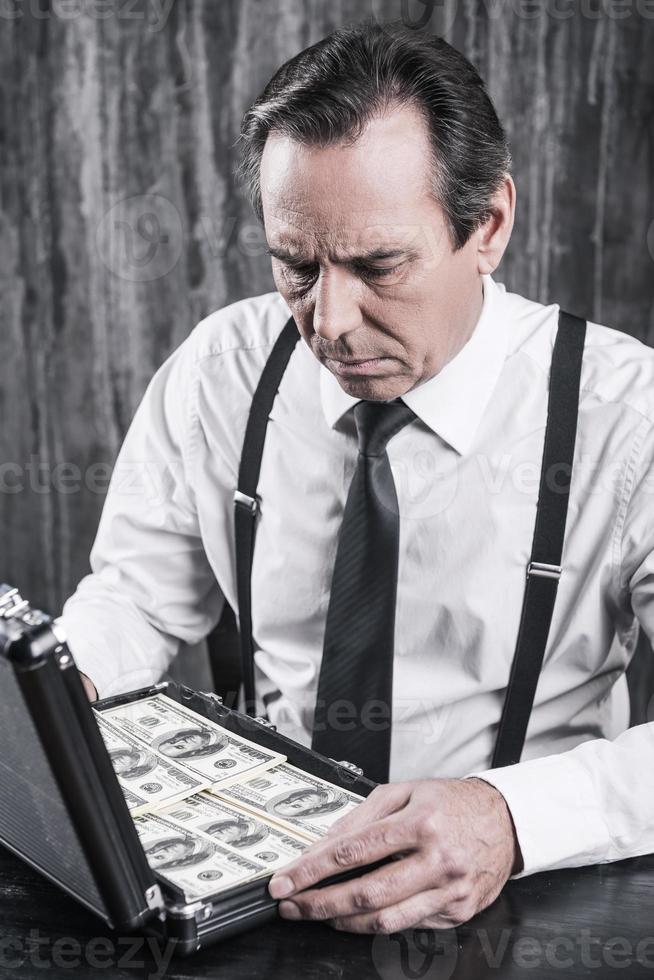 onwettig bedrijf. echt senior Mens in overhemd en bretels zittend Bij de tafel en op zoek binnen van de geval vol van papier valuta foto