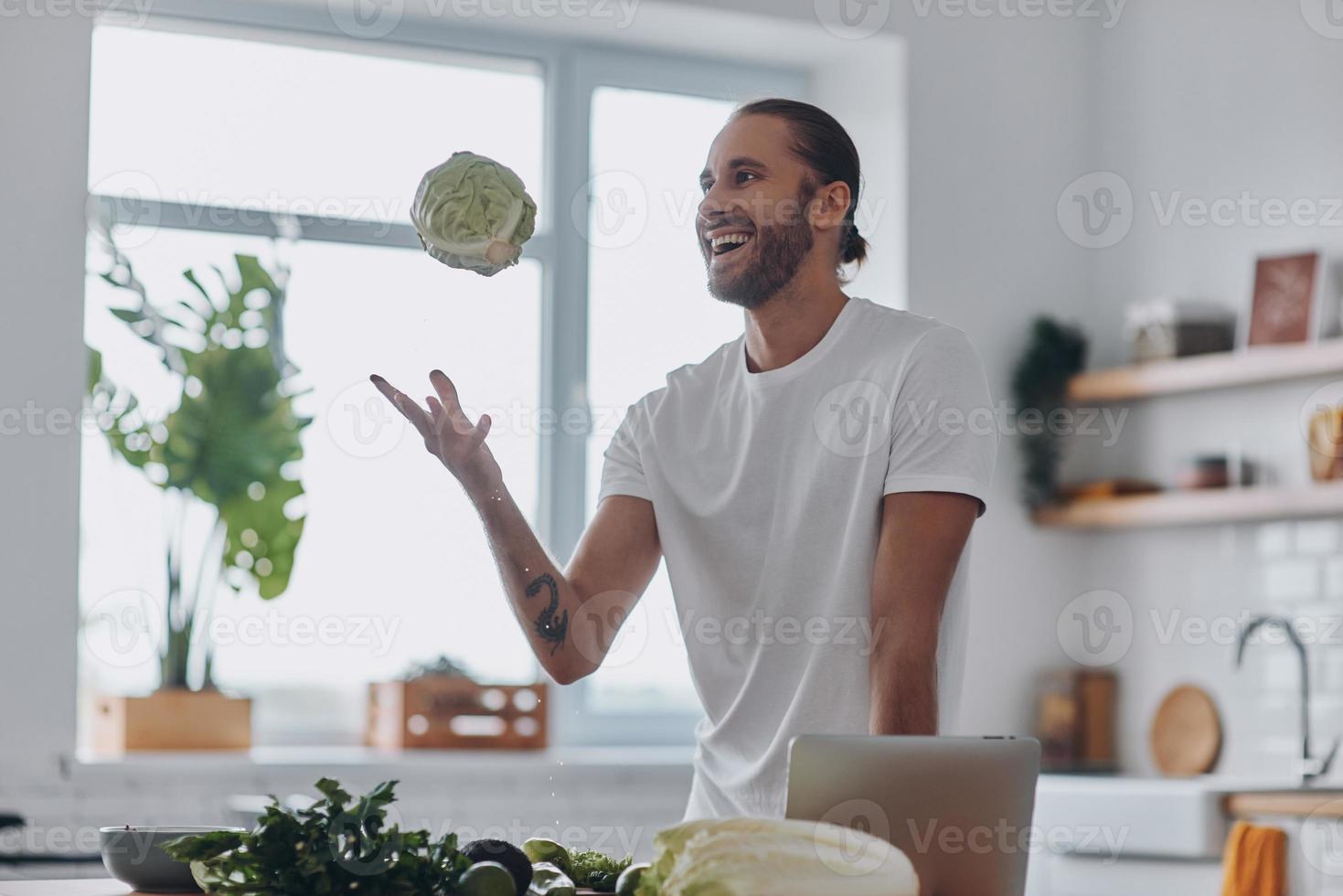 gelukkig jong Mens spelen met vers groenten terwijl Koken Bij de huiselijk keuken foto