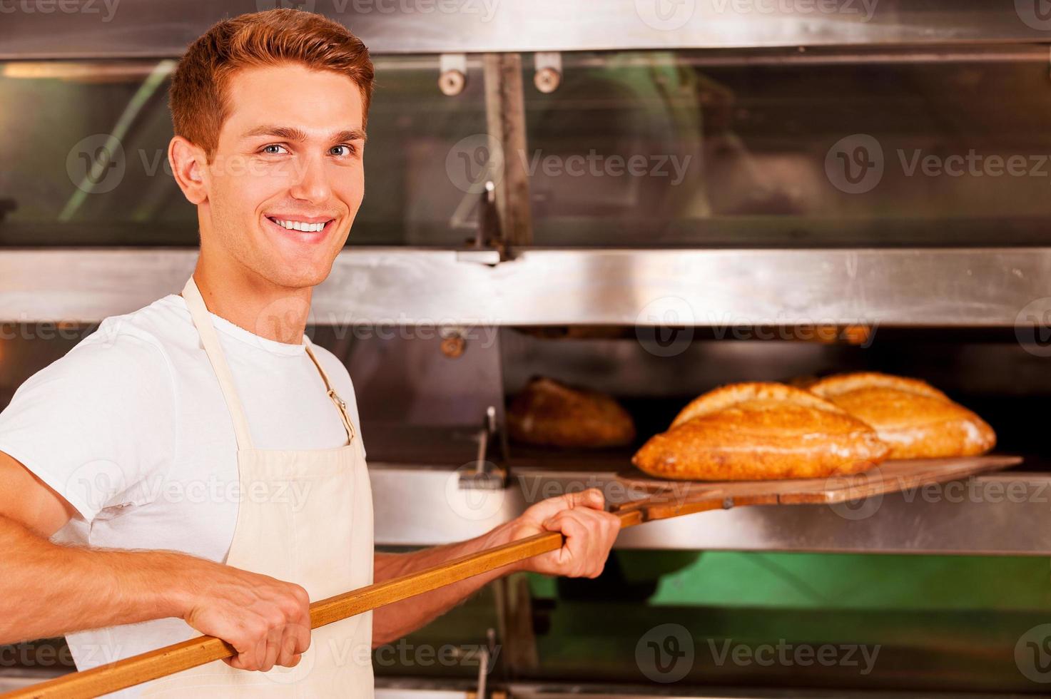 zelfverzekerd bakker. zelfverzekerd jong Mens in schort nemen vers gebakken brood van oven en glimlachen foto