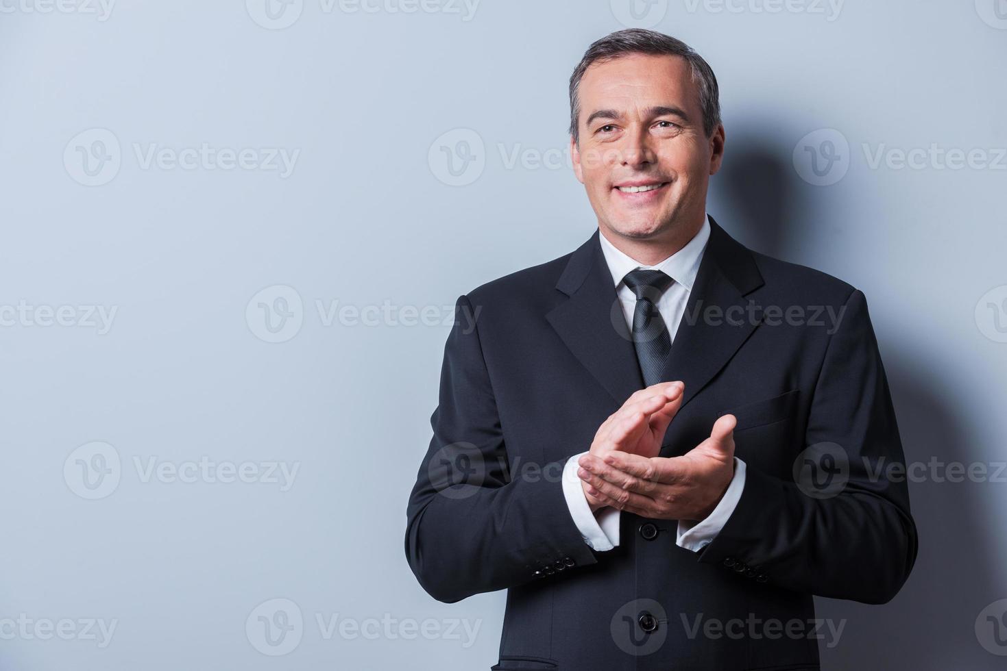 vieren succes. zelfverzekerd volwassen Mens in formele kleding klappen handen en glimlachen terwijl staand tegen grijs achtergrond foto