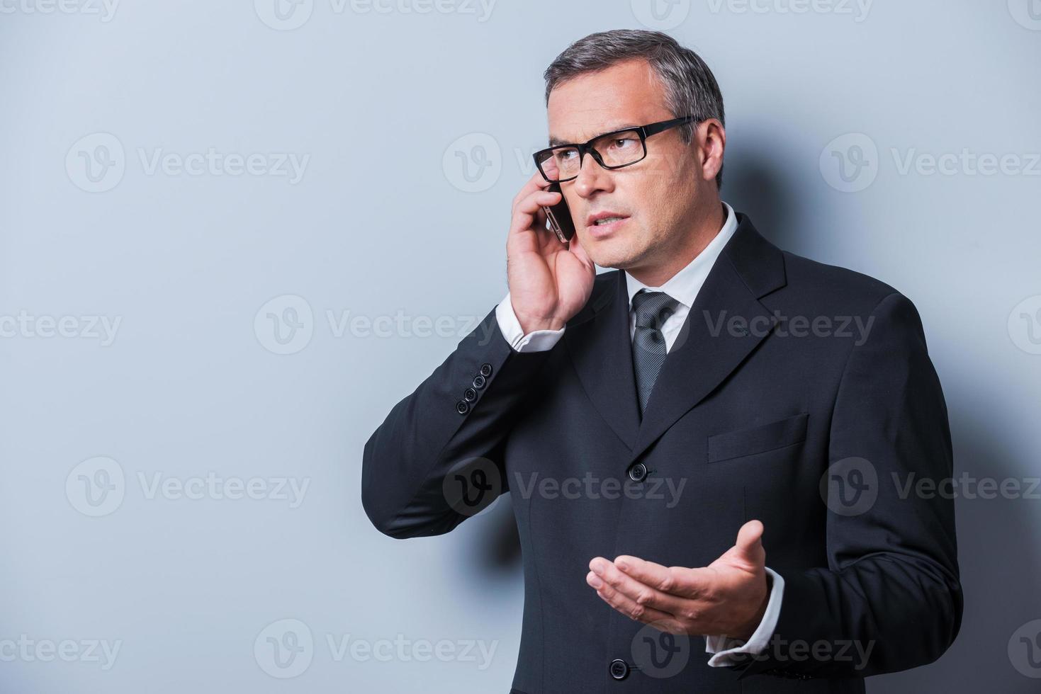 bespreken bedrijf. zelfverzekerd volwassen Mens in formele kleding pratend Aan de mobiel telefoon en gebaren terwijl staand tegen grijs achtergrond foto
