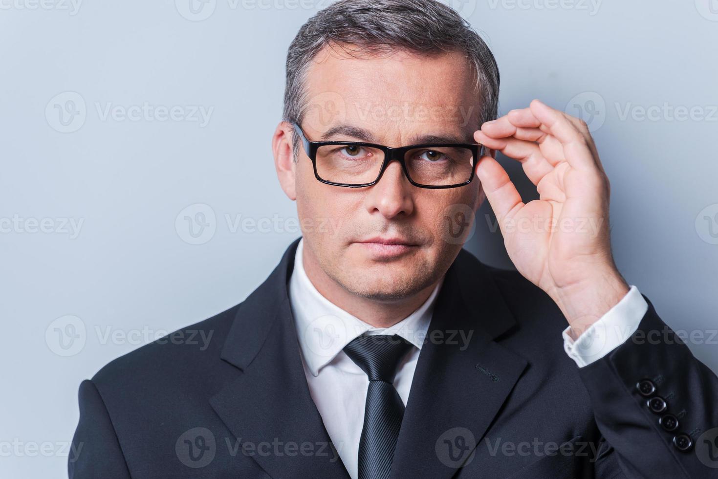 bedrijf deskundige. portret van zelfverzekerd volwassen Mens in formele kleding aanpassen zijn bril en op zoek Bij camera terwijl staand tegen grijs achtergrond foto