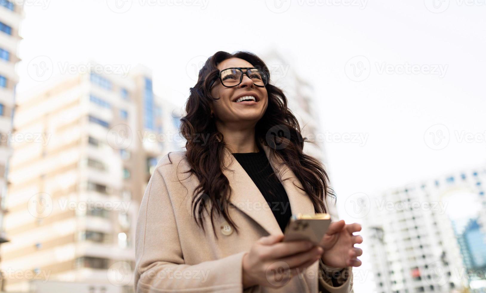 een bedrijf brunette met een mobiel telefoon in haar handen en bril voor visie tegen de backdrop van stad gebouwen met een breed glimlach foto