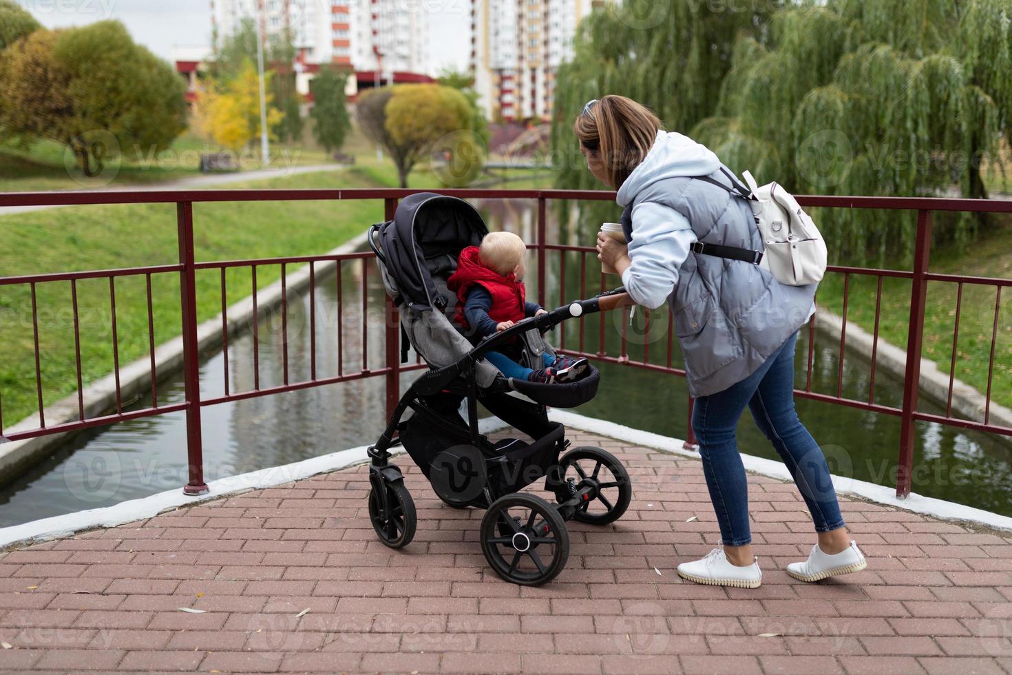een jong moeder met een rugzak Aan haar schouders wandelingen met haar kind in een kinderwagen in de stad park foto