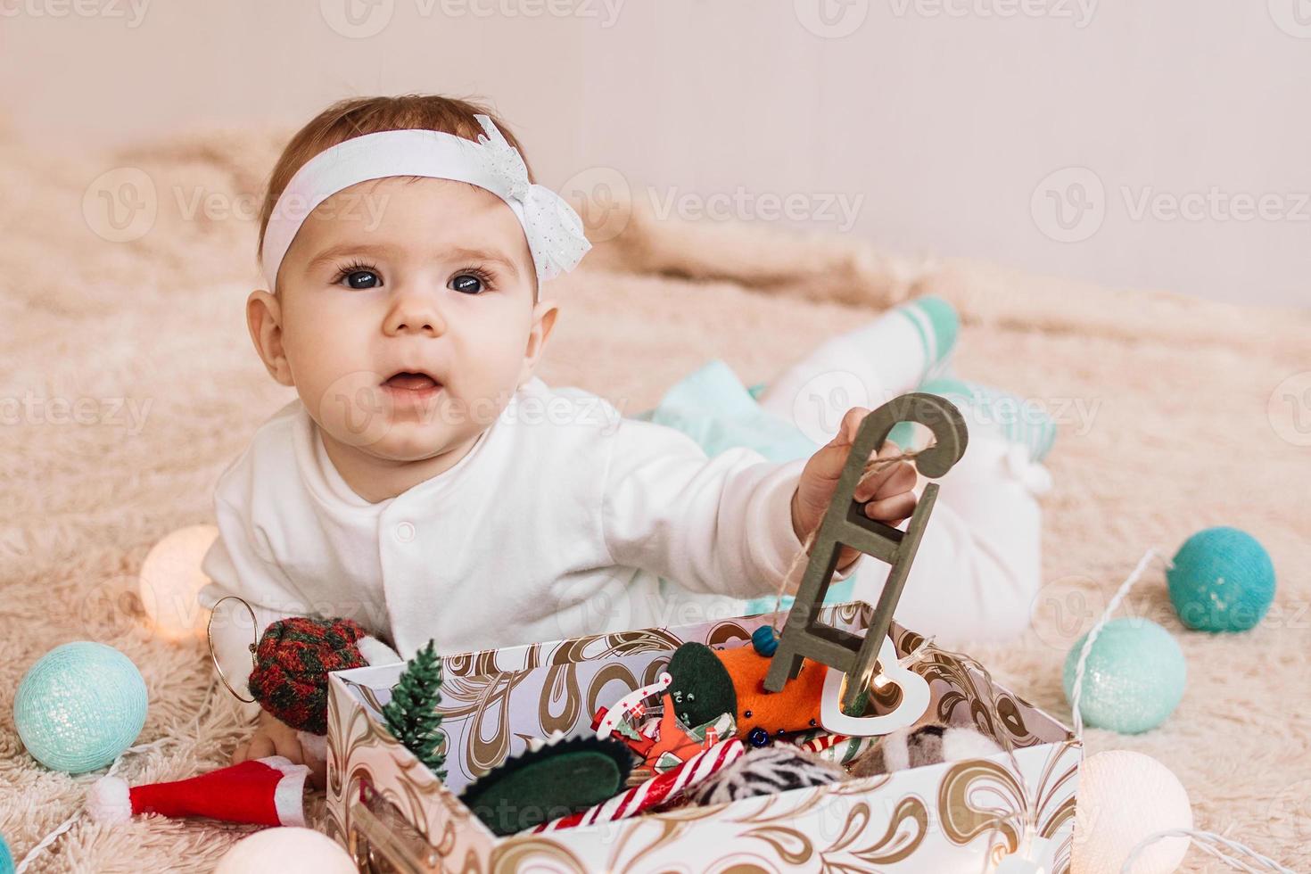 schattig weinig meisje in blauw en wit jurk is spelen met Kerstmis decoraties en Kerstmis lichten Aan beige plaid. foto