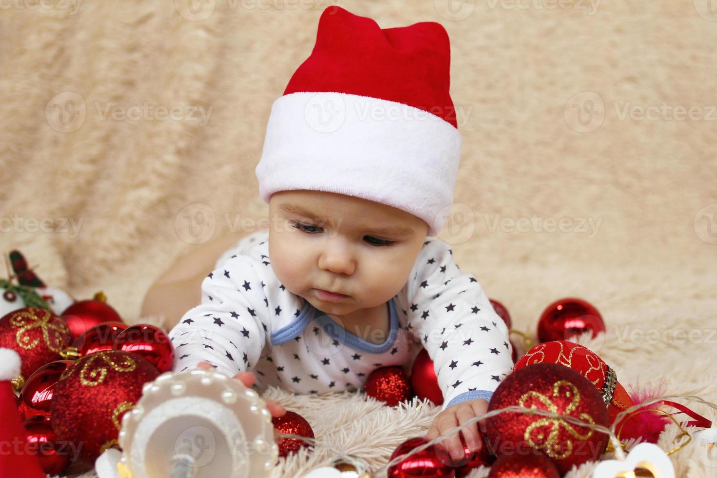 schattig weinig meisje in de kerstman claus rood hoed is spelen met rood en wit glimmend Kerstmis decoraties en Kerstmis lichten Aan een beige plaid. foto