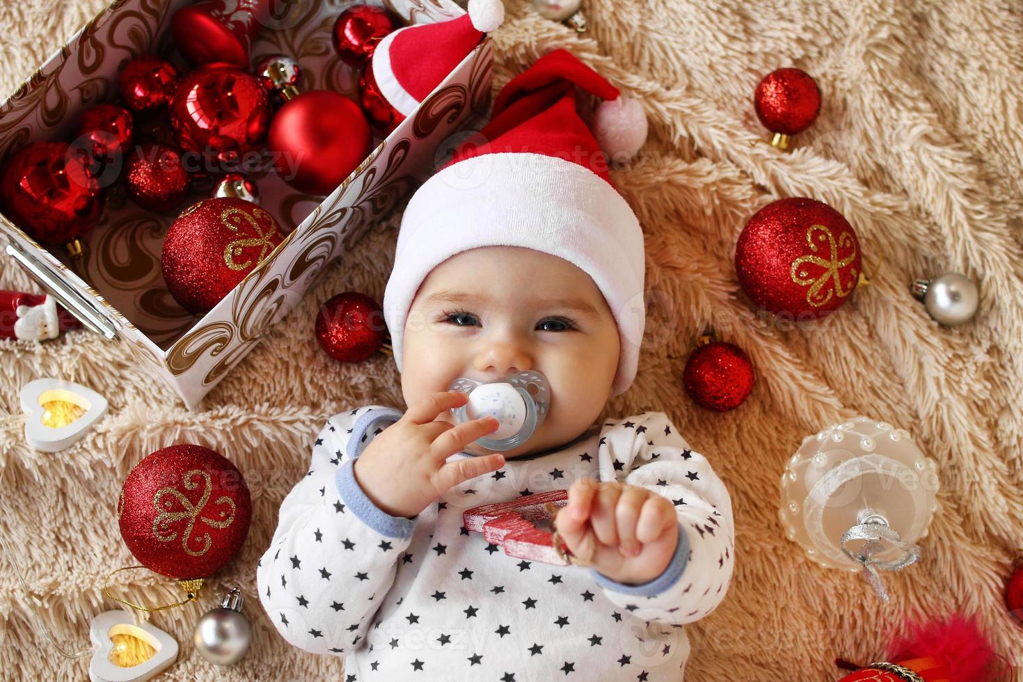 portret van schattig weinig meisje in de kerstman claus rood hoed met fopspeen Aan een beige plaid met rood en wit Kerstmis decoraties en Kerstmis lichten, top visie. foto
