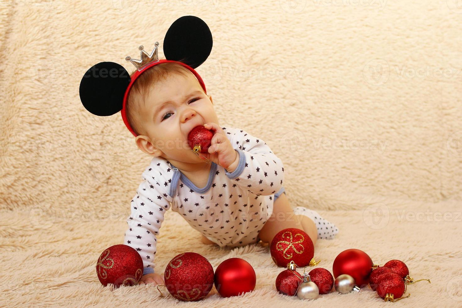 schattig weinig meisje met muis oren is zittend Aan een beige plaid en spelen met rood glimmend Kerstmis ballen. foto