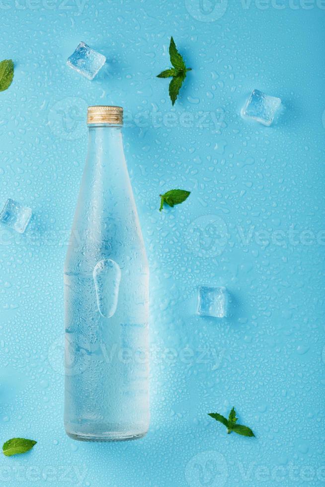 fles met een ijs verkoudheid drank, ijs kubussen, druppels en munt bladeren Aan een blauw achtergrond. foto