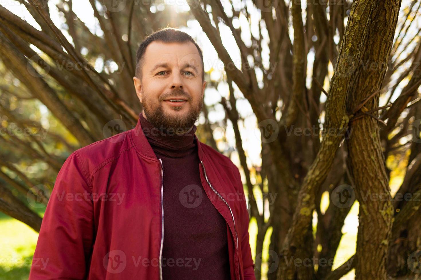 glimlachen jong Mens met baard in rood jasje in herfst park foto