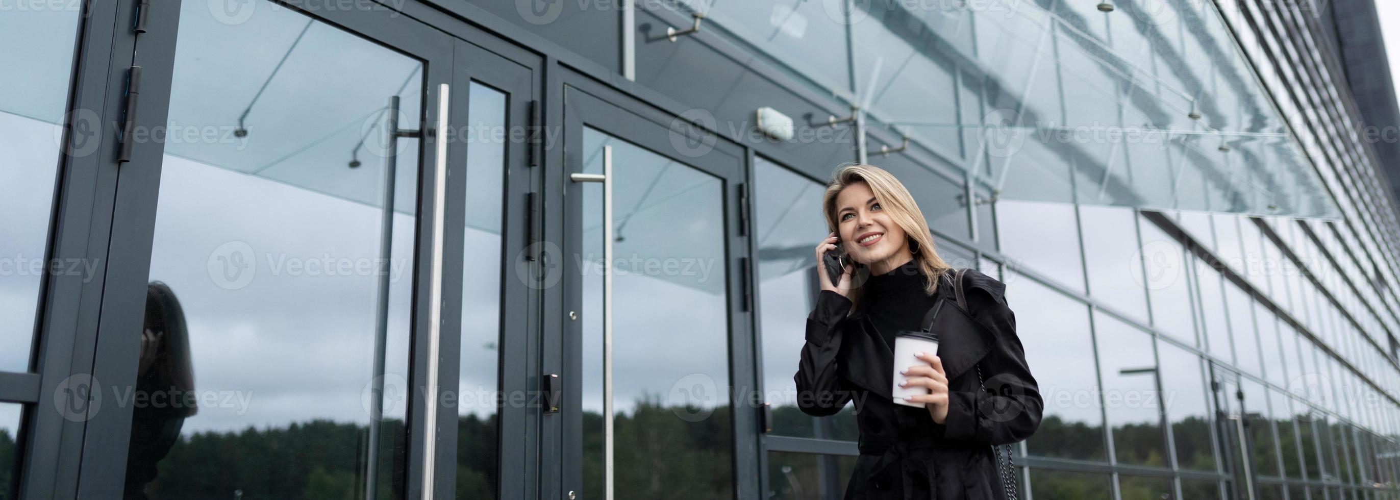 jong vrouw zakenman pratend Aan de telefoon met een glas van koffie Aan de achtergrond van een kantoor gebouw foto