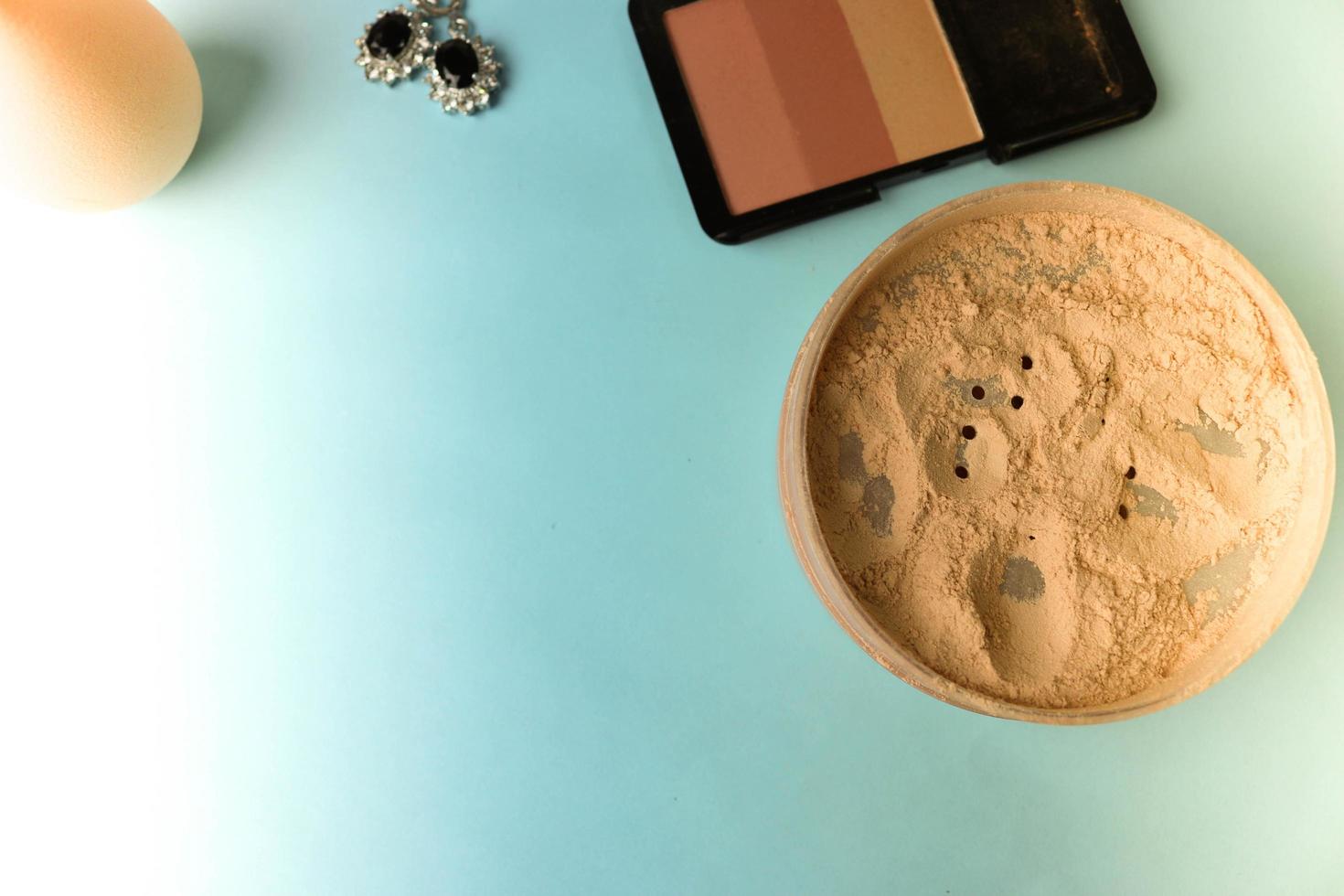 schoonheid doos, los matte mineraal poeder en blozen met een schoonheid blender voor bedenken en zilver oorbellen Aan een blauw achtergrond. vlak leggen. top visie foto