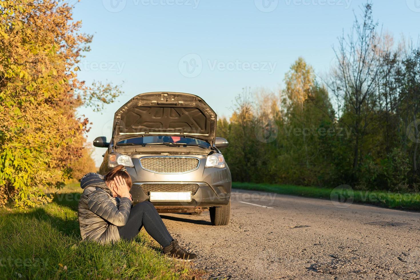 een vrouw in een jasje is proberen naar fix een gebroken auto Aan de weg. foto