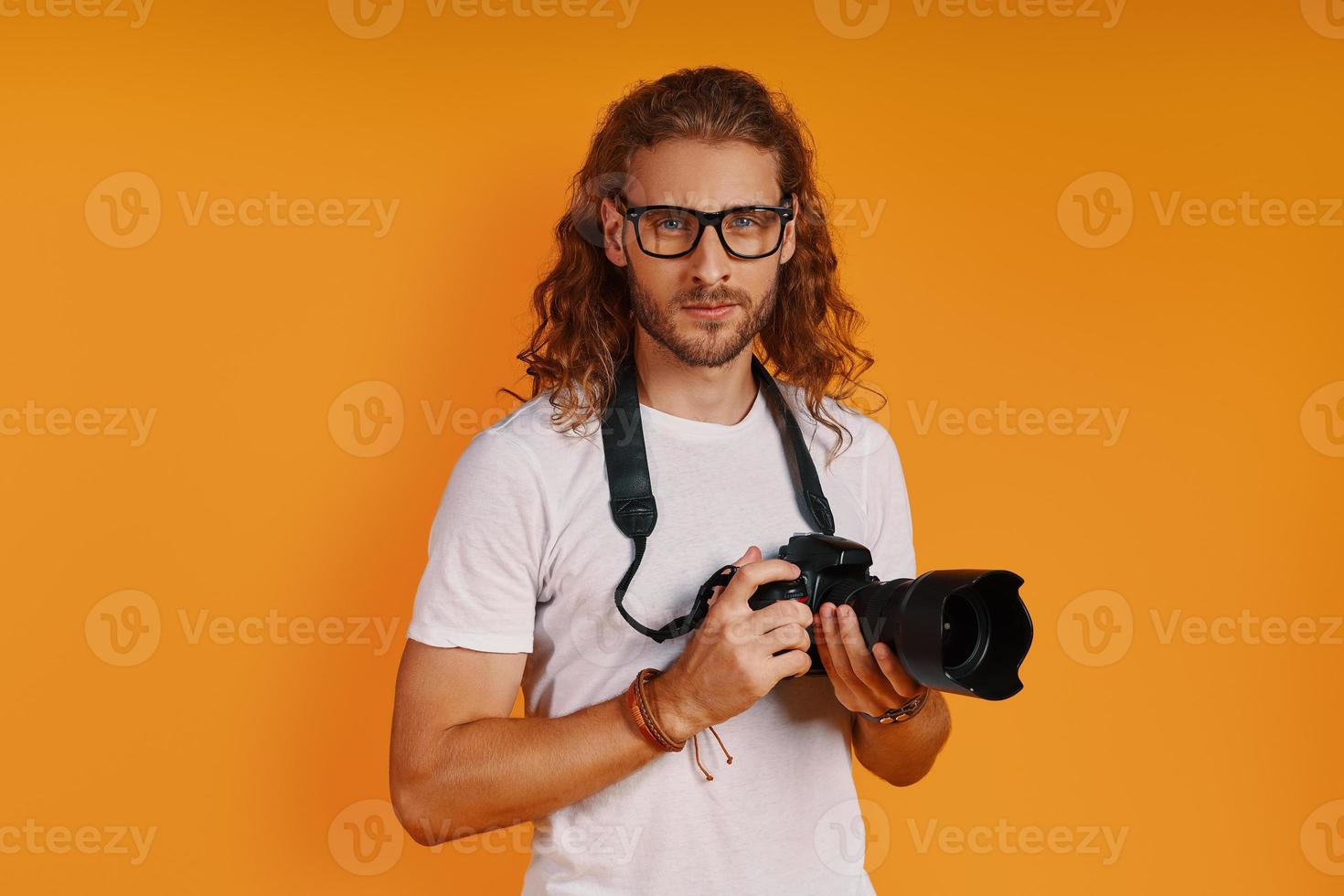 zelfverzekerd jong Mens in gewoontjes kleding op zoek Bij camera en Holding camera terwijl staand tegen geel achtergrond foto