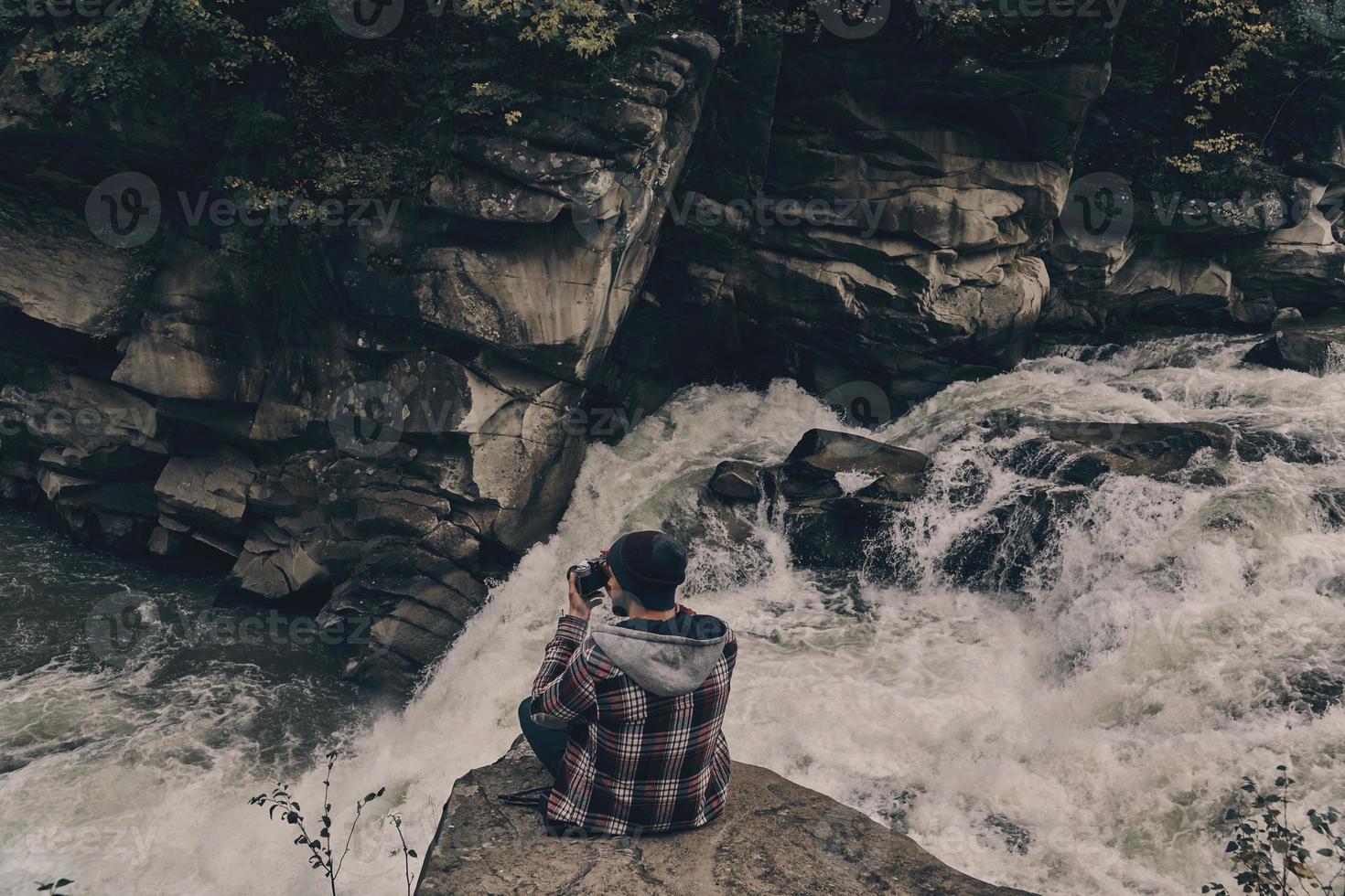 riskant functie. top visie van jong modern Mens fotograferen terwijl zittend Aan de rots met de rivier- hieronder foto